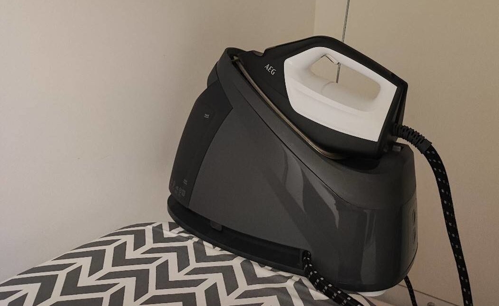 Schwarz-weiße getestete Dampfbügelstation von AEG inklusive Kabel-Halterung steht auf einer Bügelbrett-Bügeleisenablage vor weißer Wand.