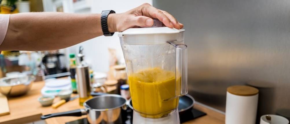 Standmixer für Suppen getestet: Eine Hand hält den weißen Deckel eines Mixbehälters fest, in dem sich eine Kürbissuppe befindet. Der Mixer steht auf einer Arbeitsfläche mit verschiedenen Utensilien.