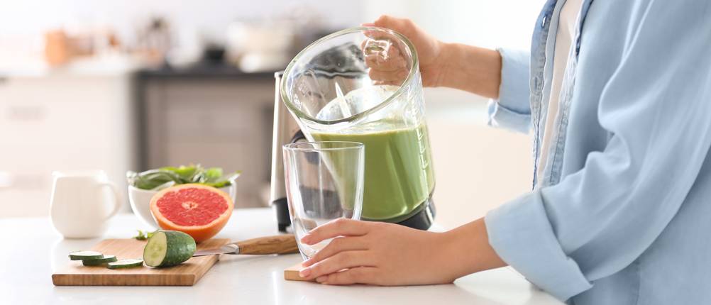 AEG-Standmixer im Test: Frau gießt mit dem Behälter eines Standmixers einen grünen Smoothie in ein Glas. Das Glas steht auf einer weißen Ablagefläche, daneben befinden sich Obst und Gemüse.