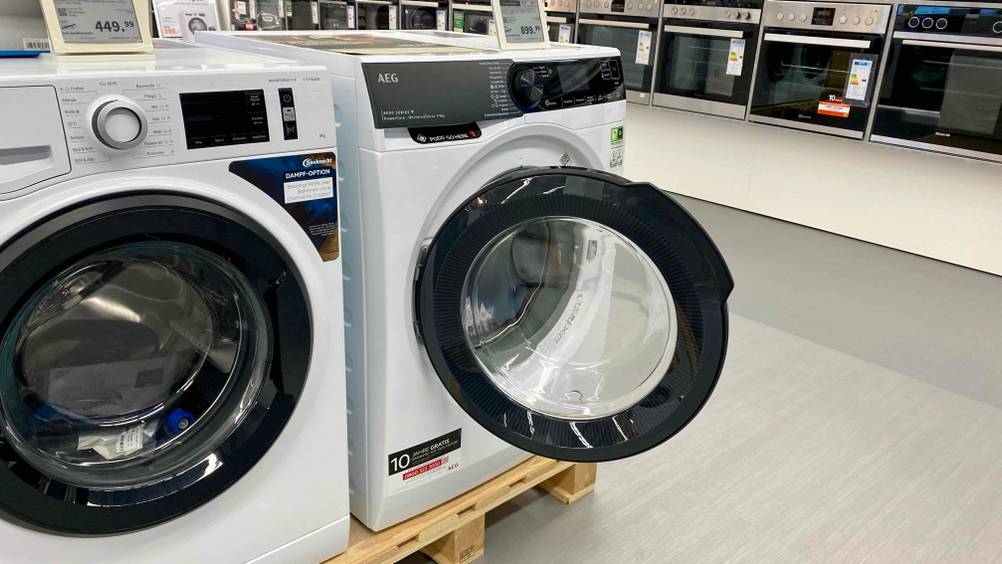 AEG-Waschmaschine getestet: Eine Maschine mit geöffneter Klappe auf einem Sockel in einer Verkaufsfläche im Hintergrund andere Geräte.