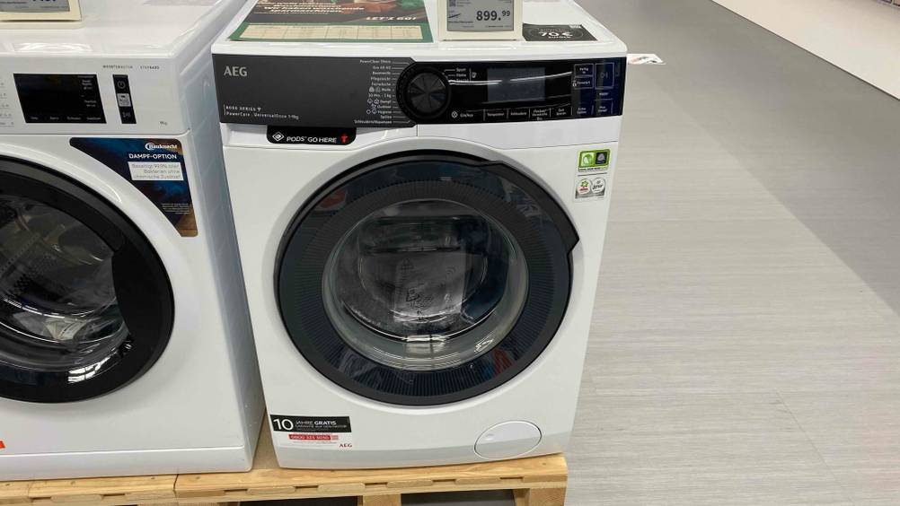 Eine getestete AEG-Waschmaschine auf einem Sockel in einer Verkaufsfläche neben anderen Produkten.