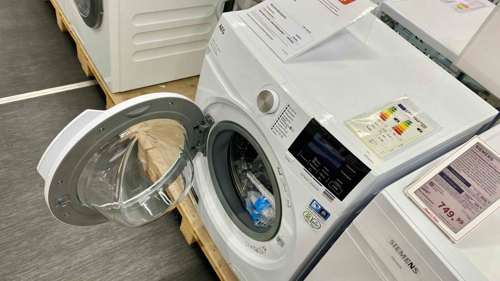 Im Test: Ein AEG-Waschtrockner von schräg oben mit geöffneter Klappe auf einem Sockel neben anderen Geräten.