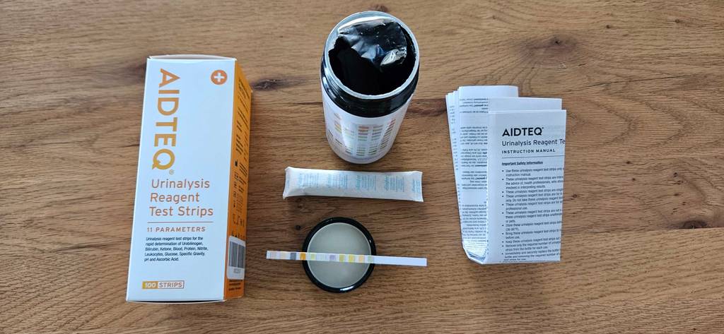 Urinteststreifen im Test: Ansicht von oben eines geöffneten, versiegelten runden Behälters, der neben dem Zubehör, einem Urinteststreifen und seiner Verpackung auf einer Holzoberfläche steht.