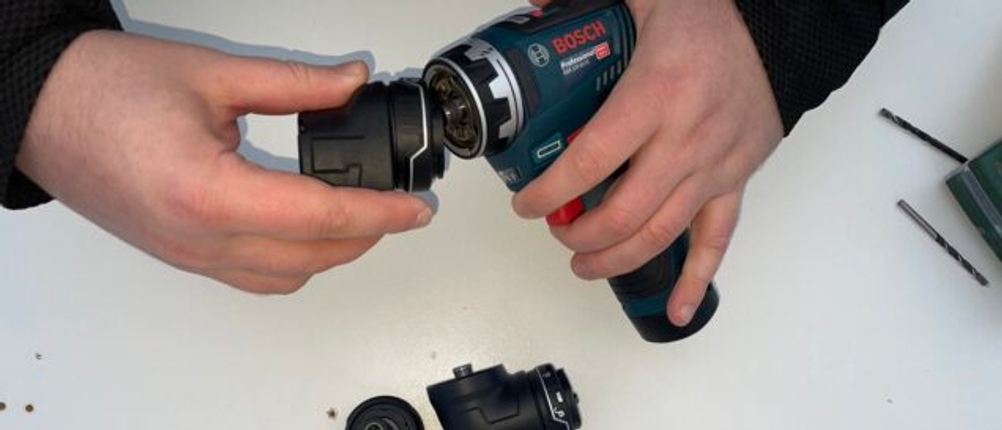 Der Bosch GSR 12V-35 FC wird mit beiden Händen gehalten und zeigt den werkzeuglosen Wechsel der FlexiClick-Aufsätze. Auf hellem Untergrund liegen weitere Aufsatzmodule und Zubehörteile des Systems.