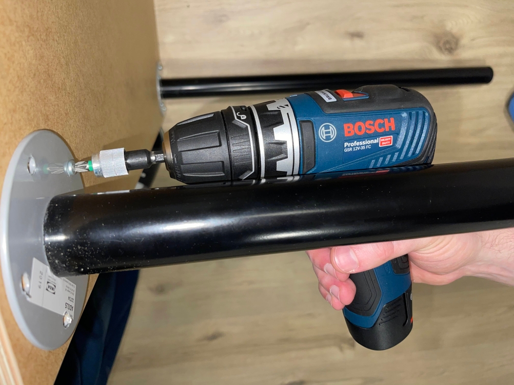 Der Bosch GSR 12V-35 FC bohrt in eine Kunststoffhalterung an einer hellen Wand. Das kompakte Gerät wird sicher mit einer Hand geführt, während der Bohrer präzise ansetzt.