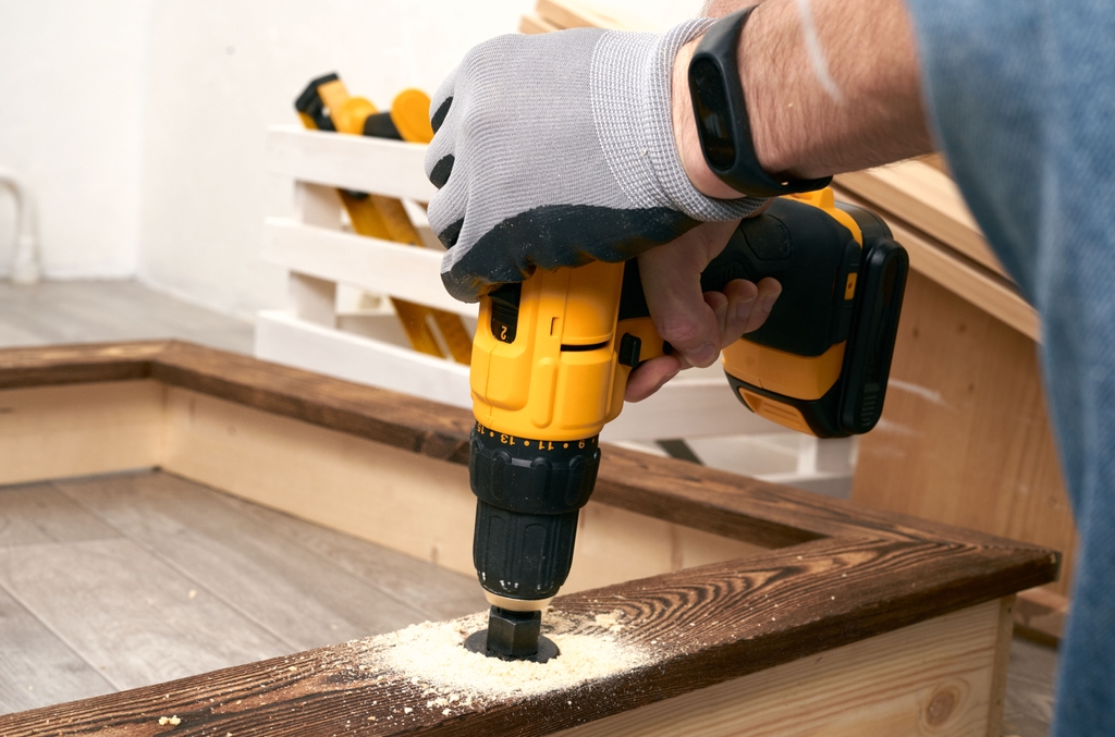 Ein Akku-Schlagbohrschrauber bohrt mit einer Lochsäge in ein Holzbrett. Die Hand hält das Werkzeug während Holzspäne auf der Oberfläche sichtbar sind.