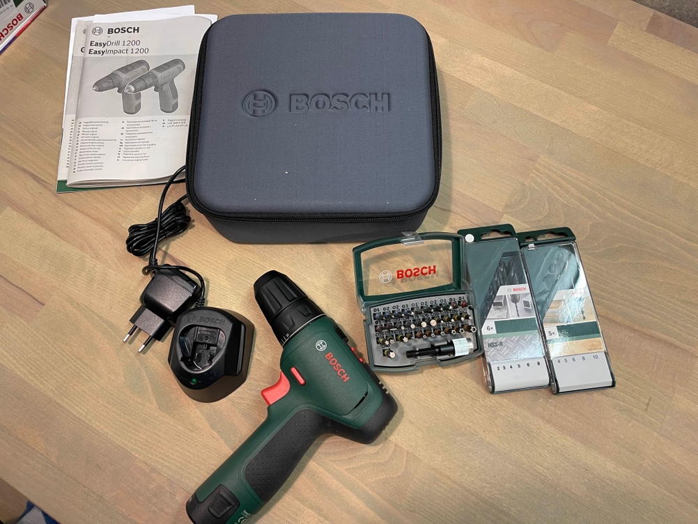 Der Bosch EasyDrill 1200 liegt ausgepackt auf einer Holzfläche, umgeben von Transportkoffer, Ladegerät, Akku, Bedienungsanleitung und zwei Bit-Sets. Der vollständige Lieferumfang zeigt alle Komponenten für den sofortigen Einsatz.