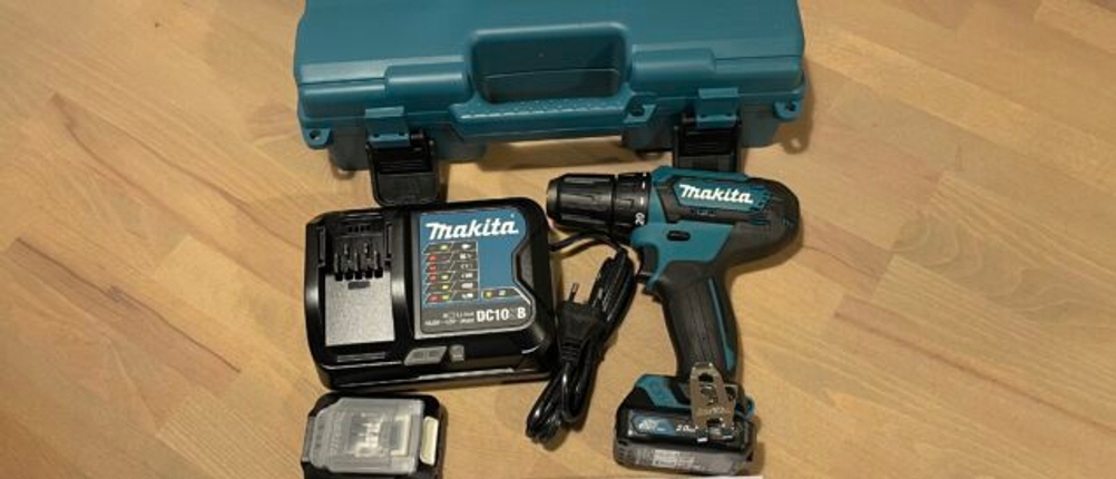 Makita DF333DSAE Akkuschrauber mit Akku, Ladegerät und Zubehör liegt ausgebreitet auf einem Tisch. Der geöffnete Transportkoffer befindet sich im Hintergrund.