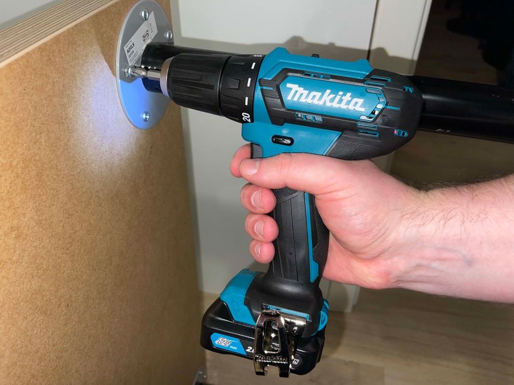 Makita DF333DSAE Akkuschrauber wird von einer Hand gehalten und setzt eine Schraube in eine Holzplatte ein. Das integrierte LED-Licht leuchtet den Schraubpunkt aus.