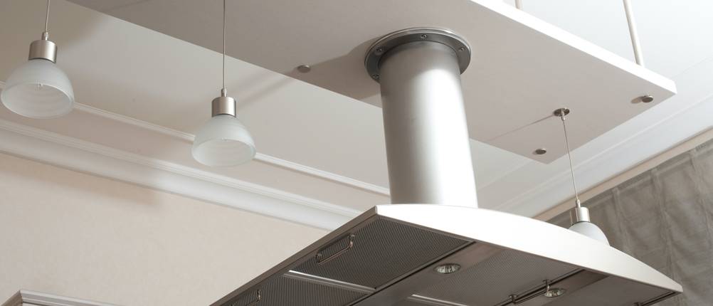 Aktivkohlefilter Dunstabzugshaube Test: Eine Inselhaube in Silber in einer sehr hohen Küche, bei der für Haube und Lampen eine extra Ebene eingezogen wurde.