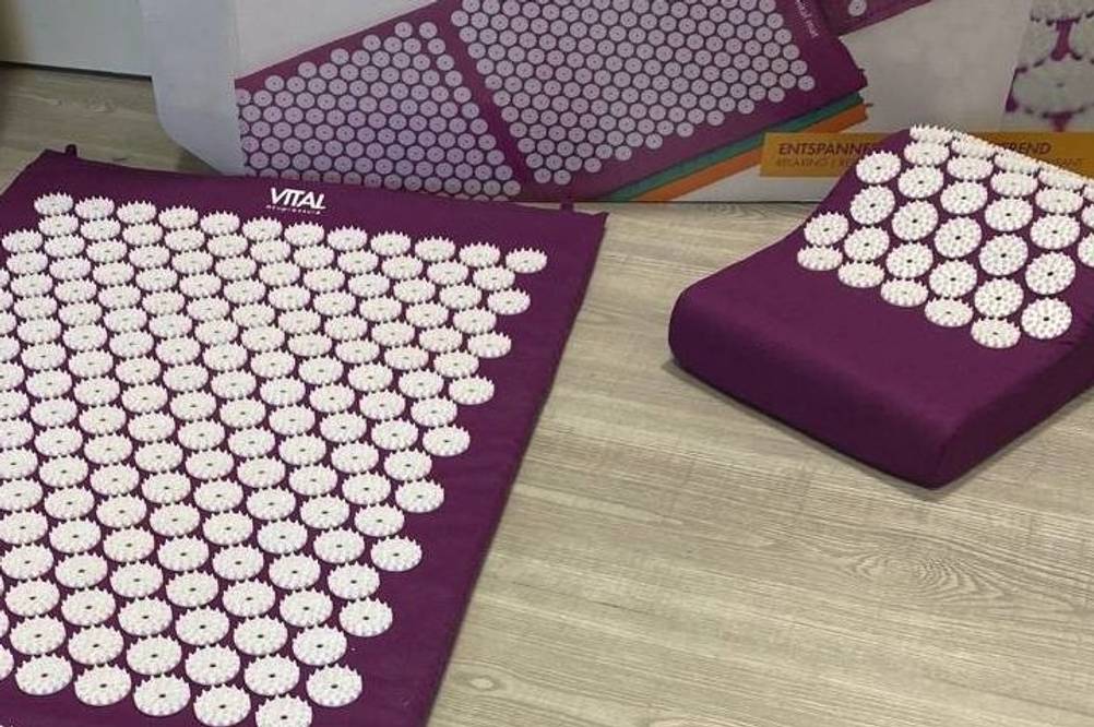 Bodhi-Akupressurmatte im Vergleich liegt aufgeschlagen auf einem Holzboden inklusive Kissen vor der Originalverpackung.