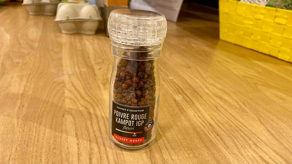 Eine Einweg-Mühle mit rotem Kampot-Pfeffer im Test steht auf einem Holztisch vor Eierpackungen.