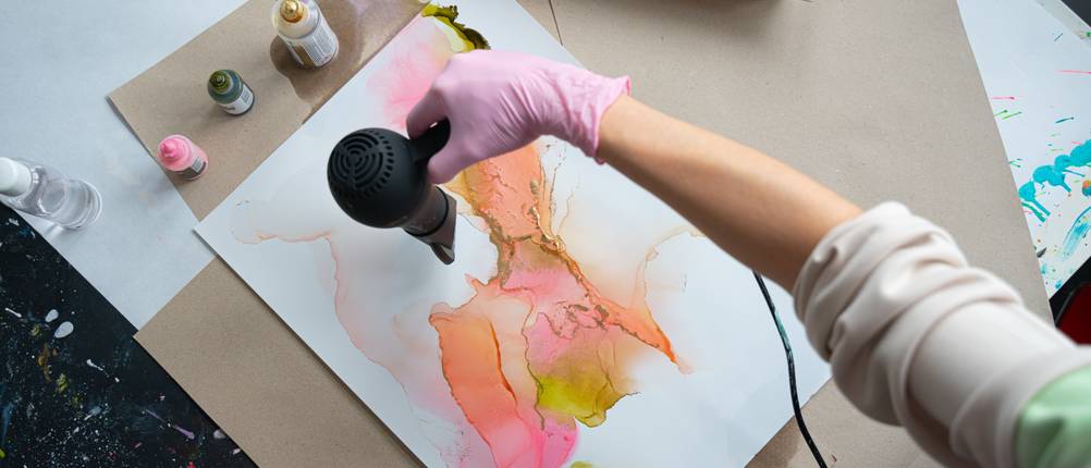 alcohol-ink-test: Frau richtet schwarzen Föhn auf ein Alcohol-Ink-Bild.