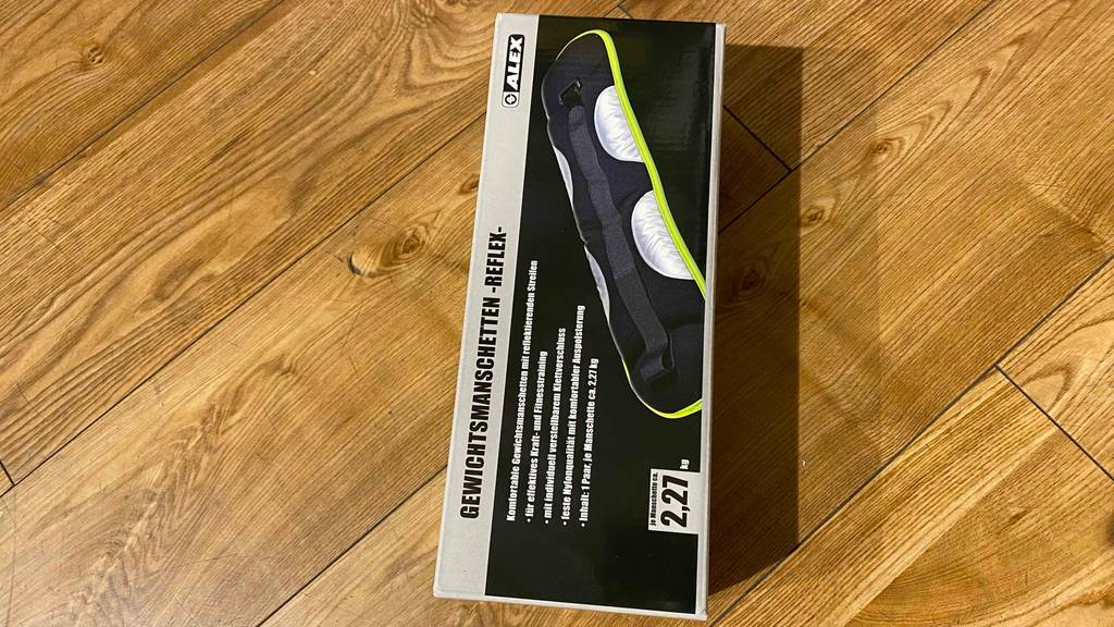 Eine Verpackung Alex Gewichtsmanschetten im Test liegt auf dem Holz-Boden.