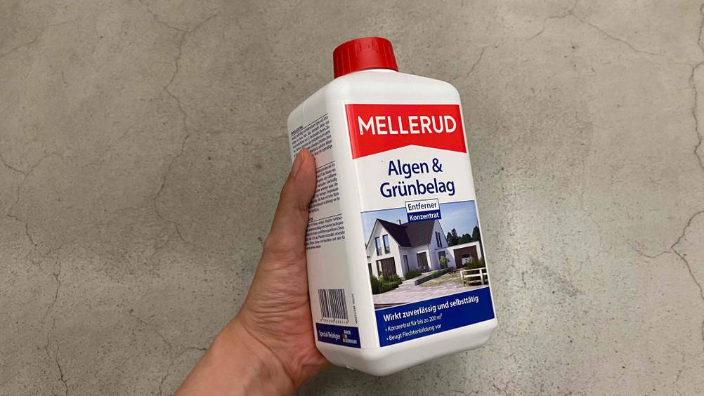 Im Test: Eine Hand hält eine Flasche Algenentferner leicht schräg vor eine Betonfläche.