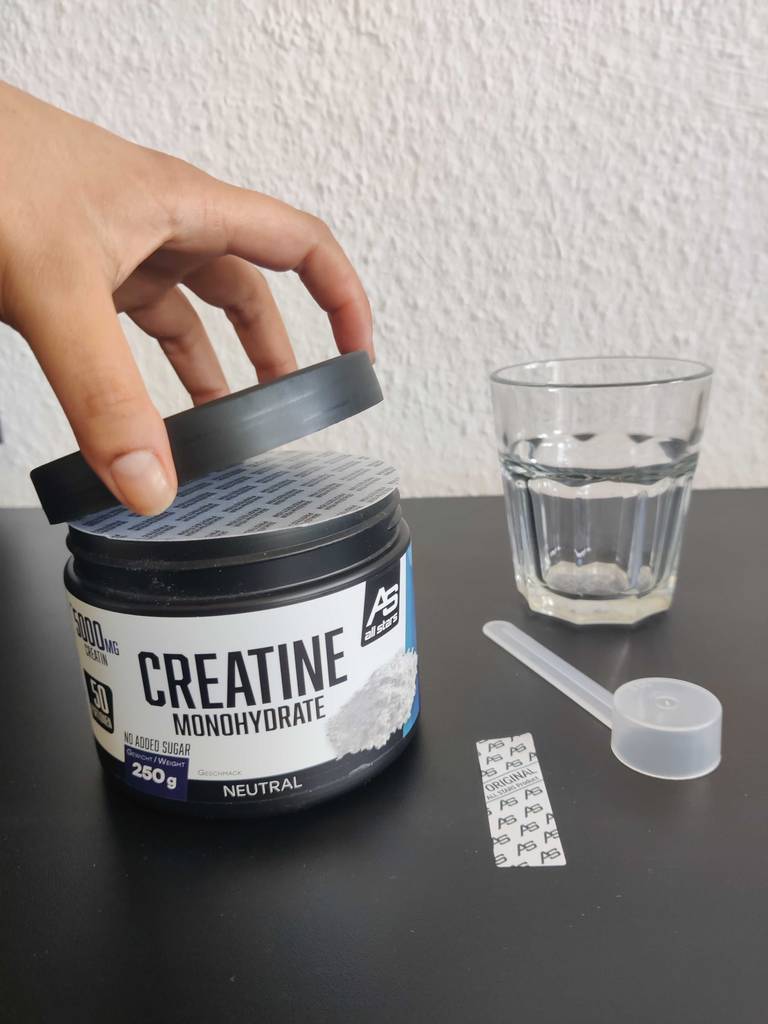 Kreatin-Pulver im Test steht in seiner Kunststoffdose neben einem Wasserglas und dem Dosierlöffel.