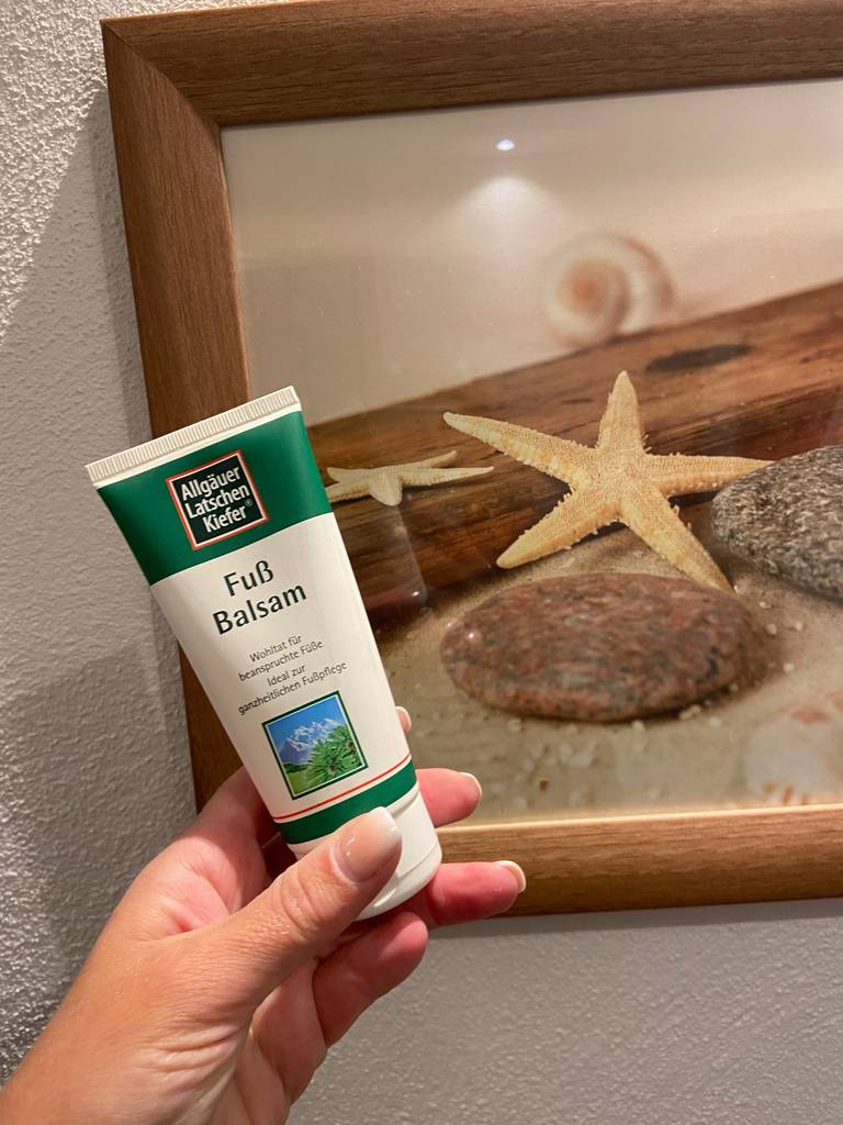 Eine Hand hält eine Tube getestete Hornhaut-Creme vor einem Sand-Dekobild.
