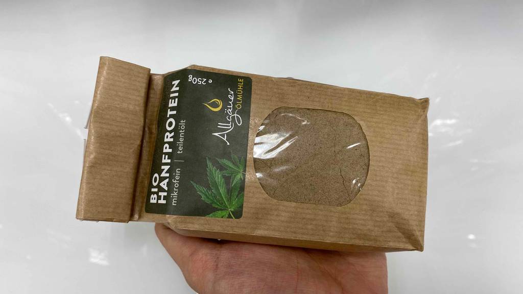 Im Test: Hand hält eine Packung Bio Hanfprotein von Allgäuer Ölmühle quer vor einem hellen Hintergrund.