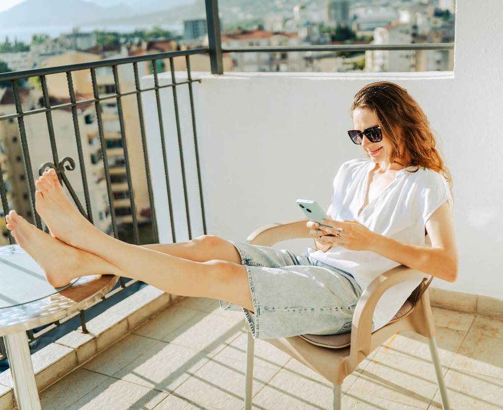 Eine junge Frau sitzt auf einem sonnigen Balkon mit ihrem Smartphone.