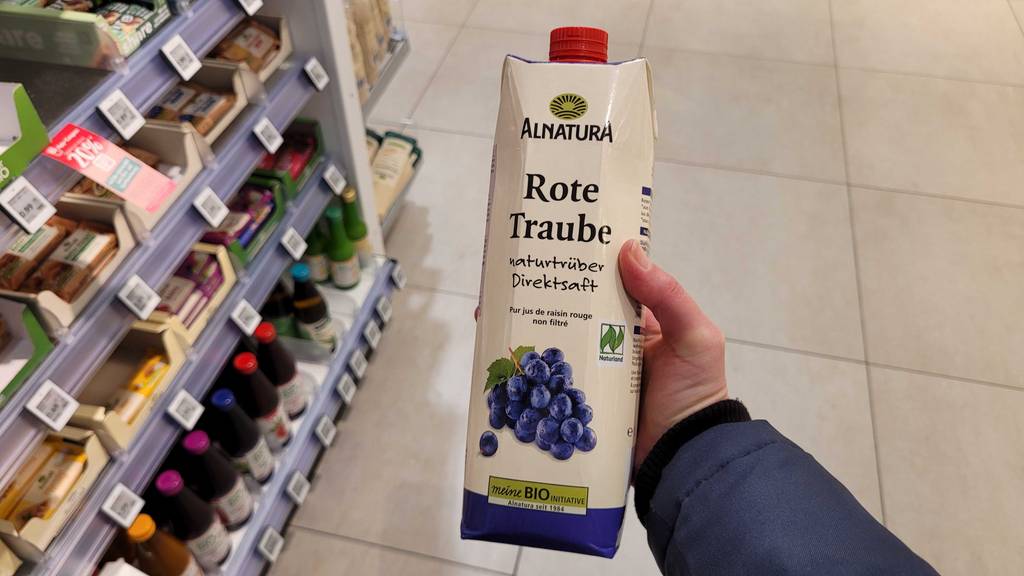 Eine Hand hält ein Tetrapak mit getestetem roten Traubensaft über dem Boden eines Drogeriemarktes.