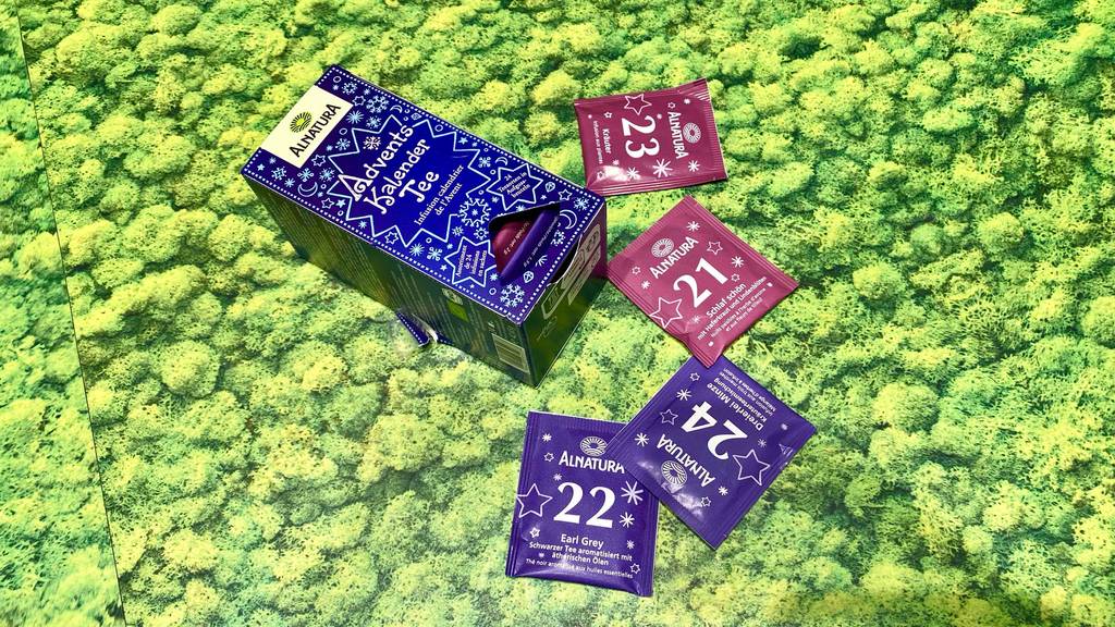 Im Test: Tee-Adventskalender der Marke Alnatura befindet sich in seiner Verpackung im blau-weißen weihnachtlichen Design und daneben liegen verschiedene einzeln verpackte Teebeutel inklusive Nummern, auf grünem Geschenkpapier.