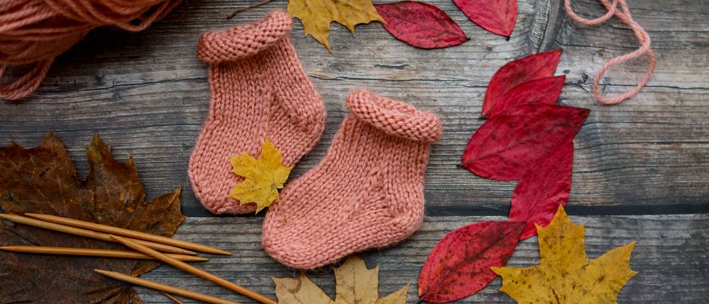 alpaka-socken-test: selbstgestrickte Babysocken aus Alpaka-Wolle mit Stricknadeln und Blättern auf Holzbretterboden