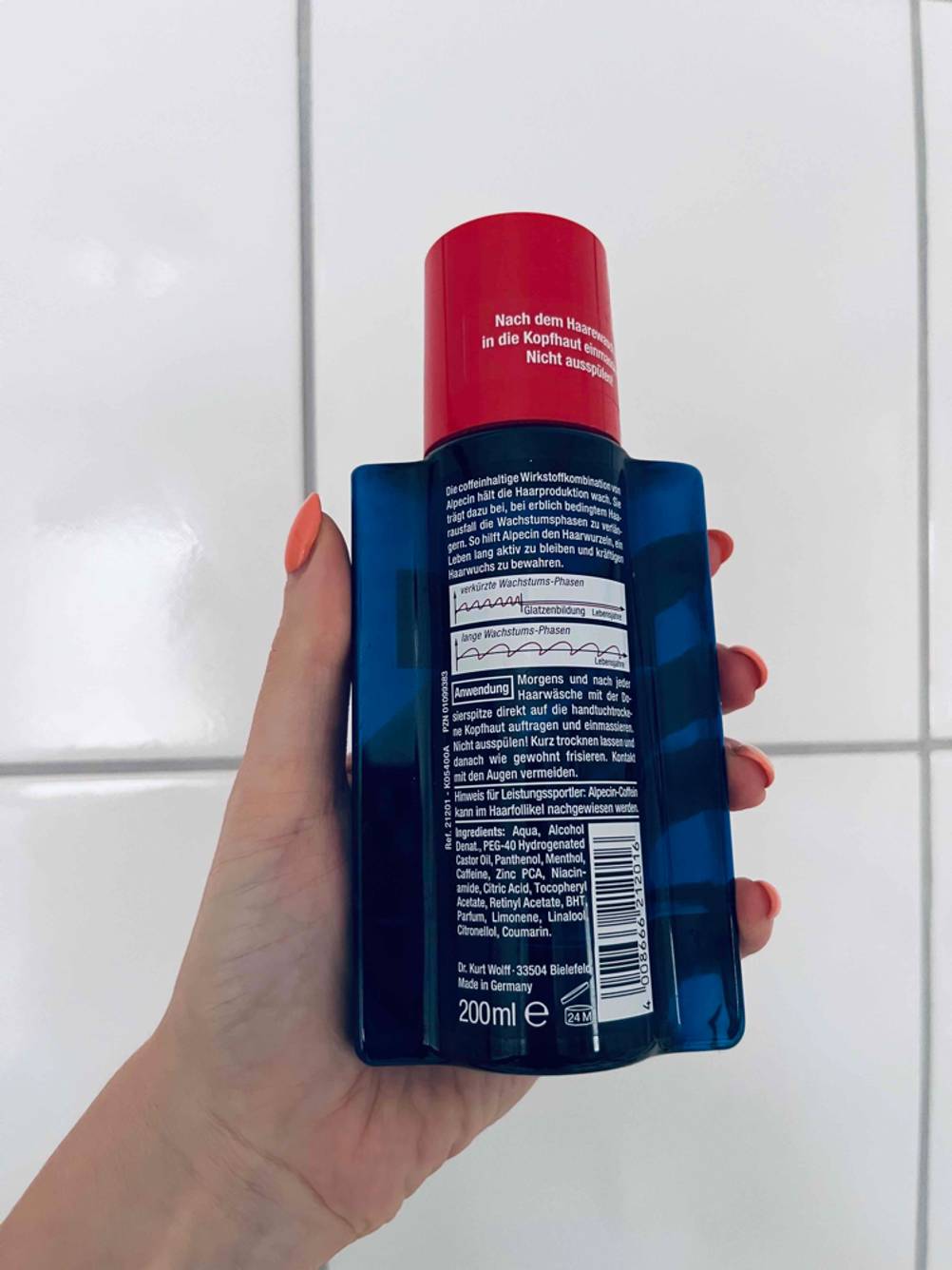 Hand hält eine 200-ml-Flasche Haarwasser Alpecin im Test, mit Fokus auf die rückseitige Anwendungsbeschreibung vor weißer Fliesenhand.