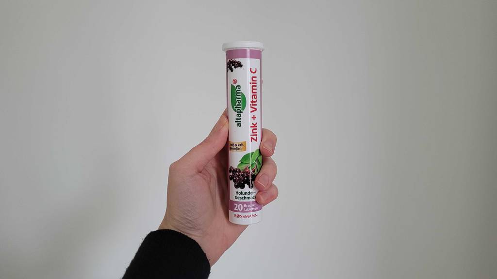 Eine Hand hält eine Dose mit getesteten Vitamin-C-+-Zink-Kapseln vor einer weißen Wand.