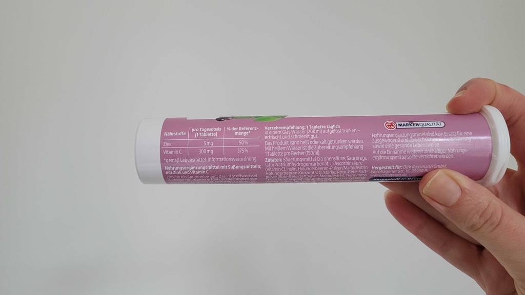 Eine Hand hält mit Fokus auf die gedruckte Verzehrempfehlung eine Dose mit getesteten Vitamin-C-+-Zink-Kapseln vor einer weißen Wand.