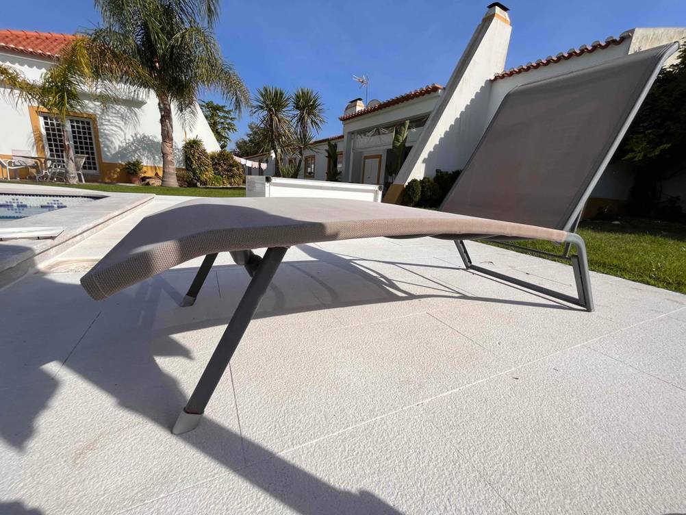 Seitenansicht einer grauen, mit Netz-Stoff bespannten und getesteten Aluminium-Sonnenliege, die am Pool in der Nähe eines Hauses steht.