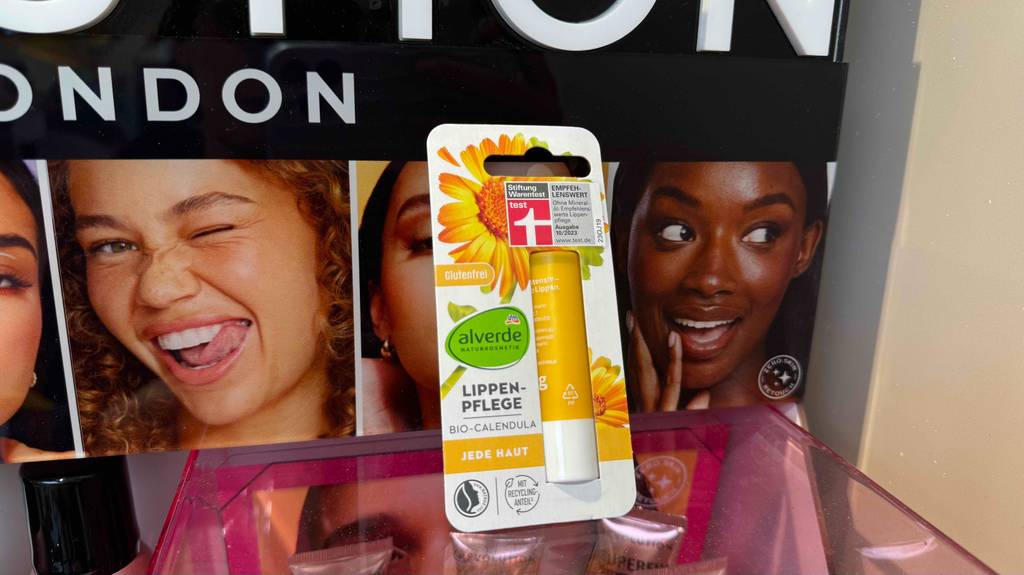 Eine getestete Lippenpflege-Naturkosmetik in der Verpackung steht vor einem Plakat mit Frauengesichtern.
