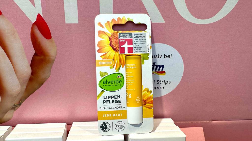 Eine Naturkosmetik-Lippenpflege im Test steht in der Verpackung vor einem Drogeriemarkt-Plakat.