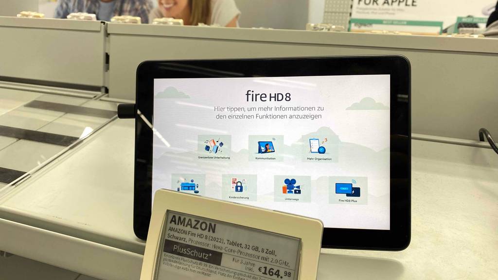 8-Zoll-Tablet-Test: ein eingeschaltetes Amazon Modell mit weißem Display, das verschiedene Programmfunktionen anzeigt, befindet sich hinter der Produktbeschreibung mit Preis auf einem weißen Ausstellungstisch.