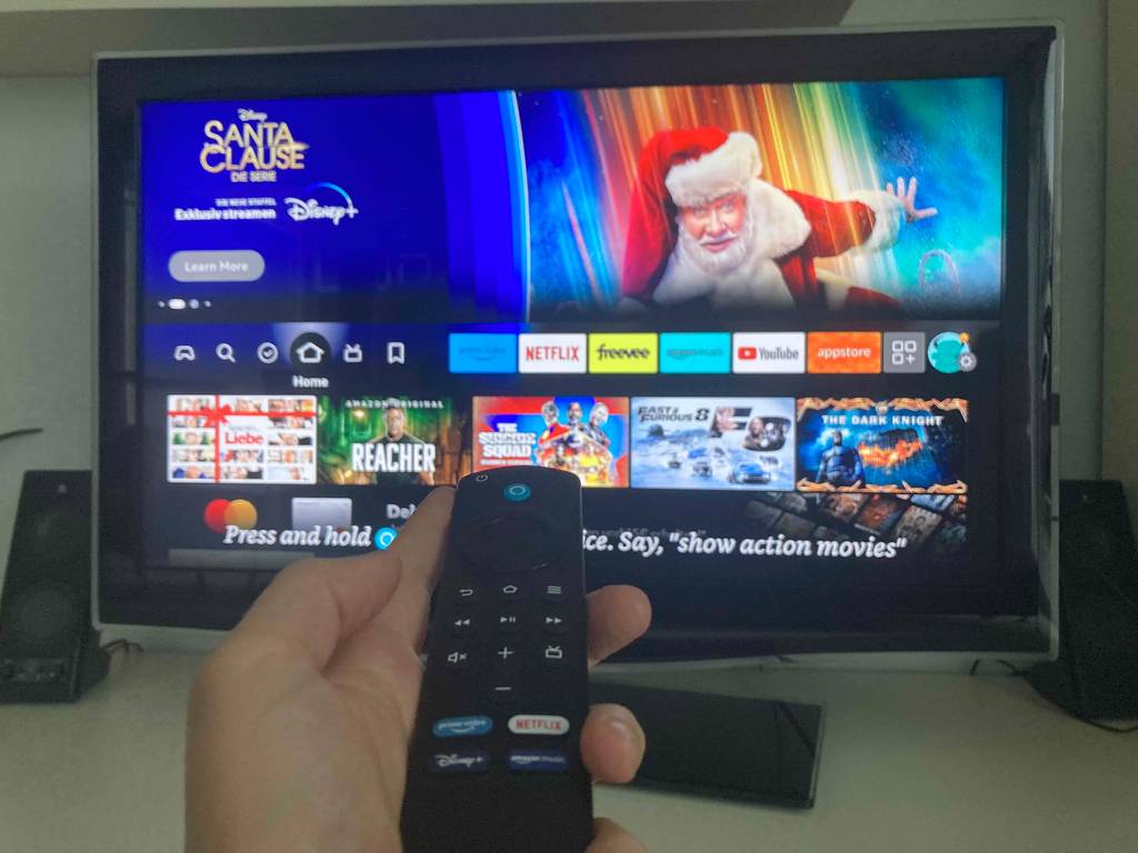 Hand hält eine Fernbedienung des getesteten Amazon-Fire-Tv-Sticks vor einen eingeschalteten Fernsehapparat.