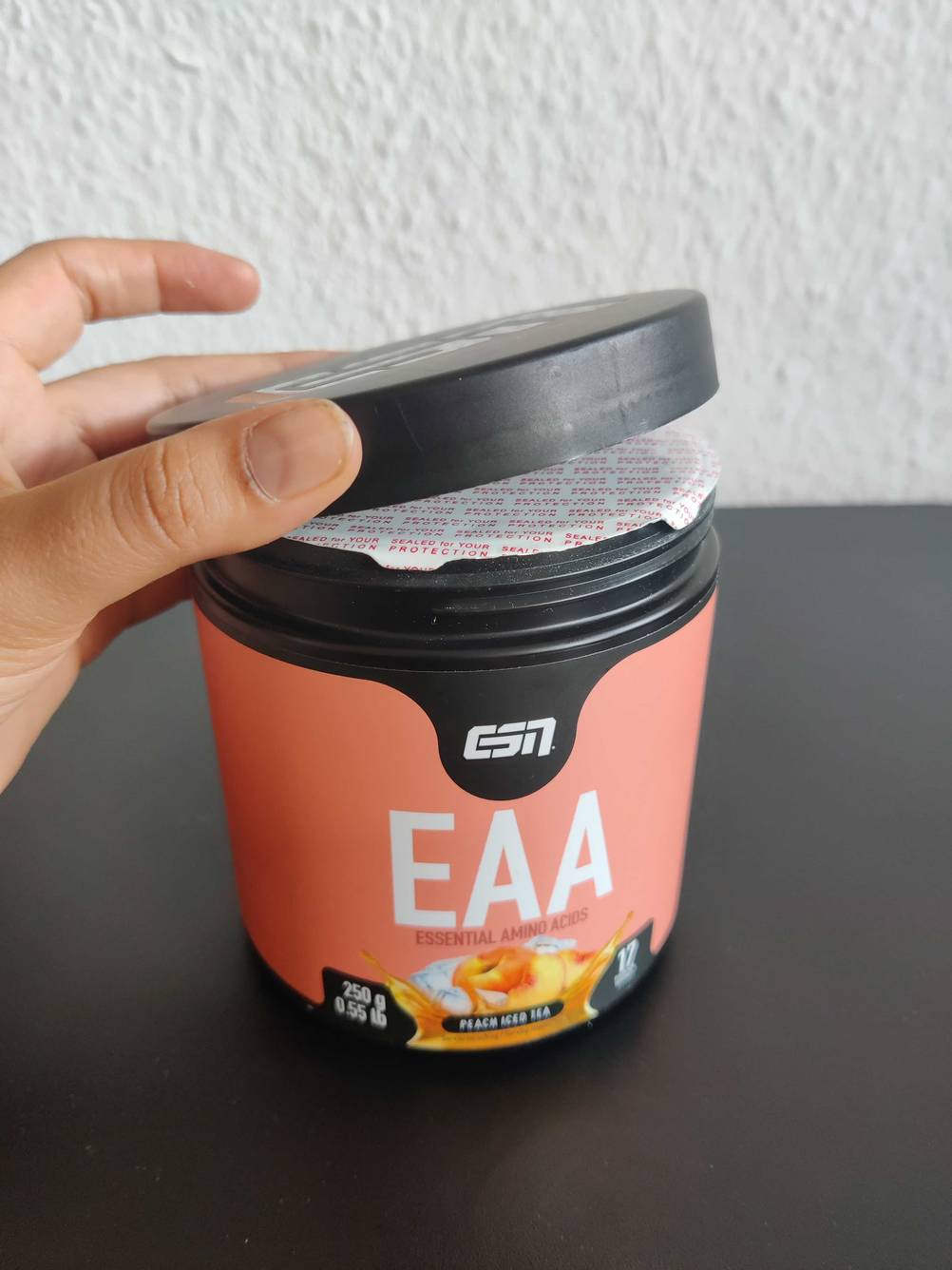 Eine Hand entfernt den Deckel einer Dose des getesteten Aminosäure-Komplexes auf einer dunklen Fläche.