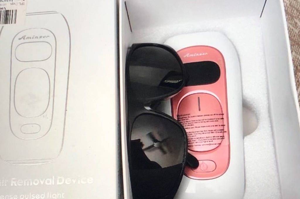 Im Test: IPL-Gerät von Aminzer in Weiß mit rosa Einstellpaneel und schwarzer LCD-Temperaturanzeige liegt in seiner weißen Verpackung, darauf befindet sich eine schwarze Sonnenbrille.