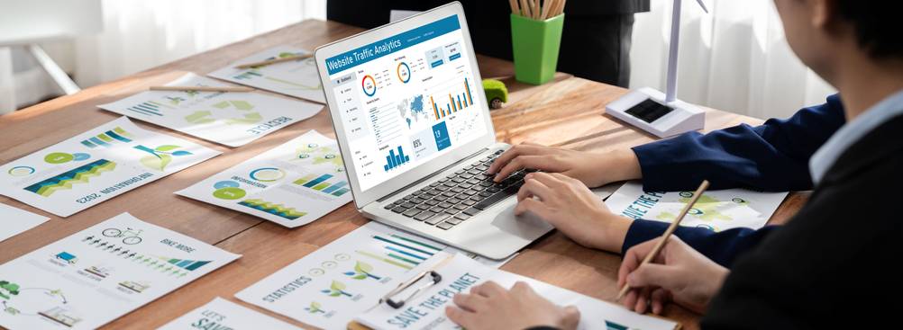 Eine Person sitzt an einem Schreibtisch auf dem Diagramme ausgebreitet sind und ein Laptop steht und analysiert den Traffic seiner Website.
