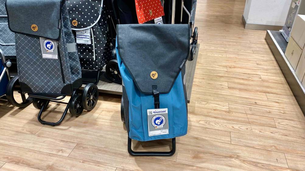 Einkaufstrolley-Test: Ein blau-grauer Trolley von vorne mit Labels auf einem Holzboden, im Hintergrund andere Produkte.