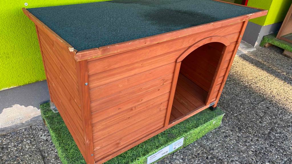 Im Test: schräge Ansicht einer Anione Hundehütte aus Holz, die auf einem Kunstrasenpodest steht.