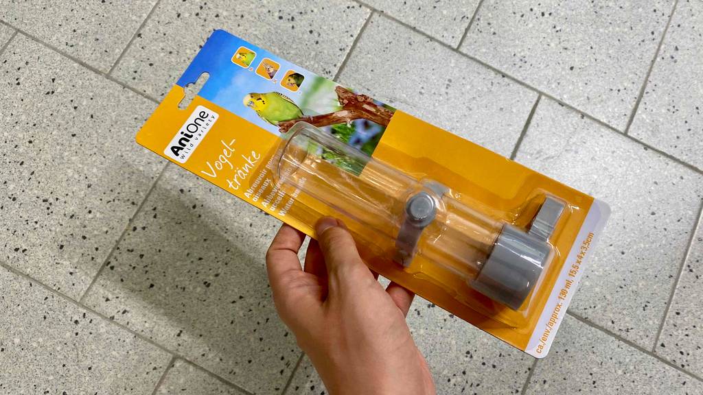 Vogeltränke im Test: Hand hält transparente Vogeltränke von Anione in seiner Verpackung vor hellgrauen Fliesen.