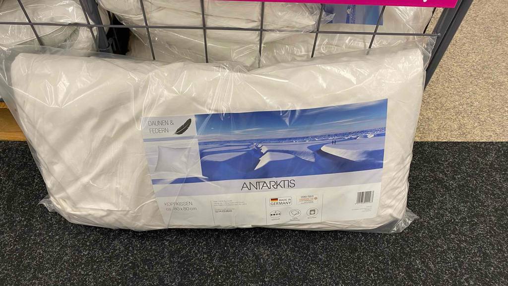 Federkissen 80x80 im Test: ein verpacktes Kissen liegt vor einer Gitterbox.