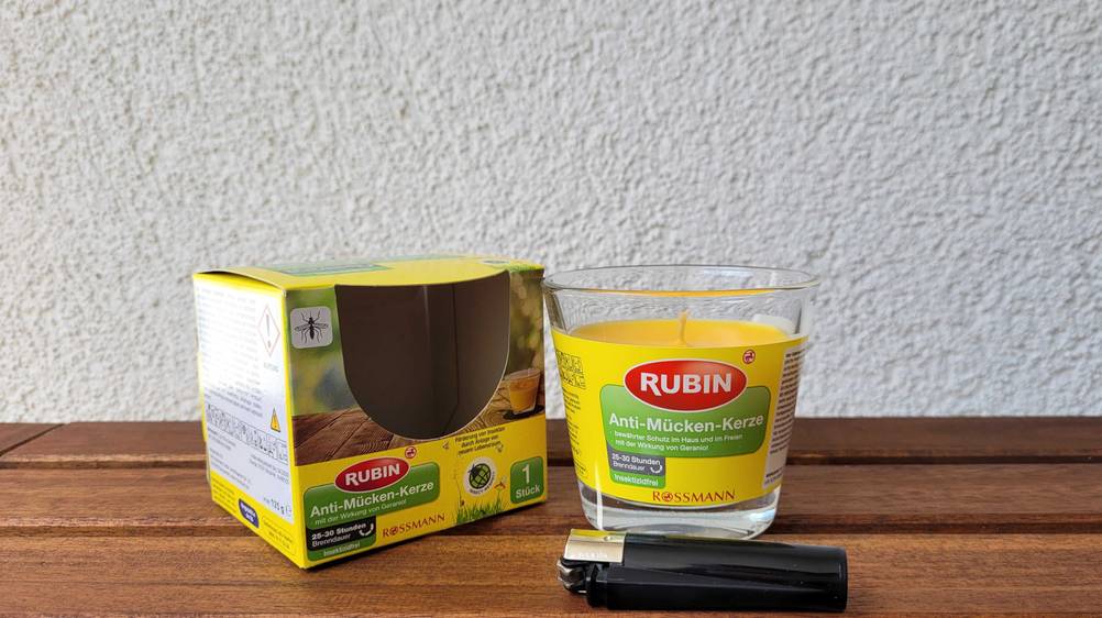 Die Anti-Mücken-Kerze von Rubin steht neben der Verpackung davor liegt ein schwarzes Feuerzeug. 