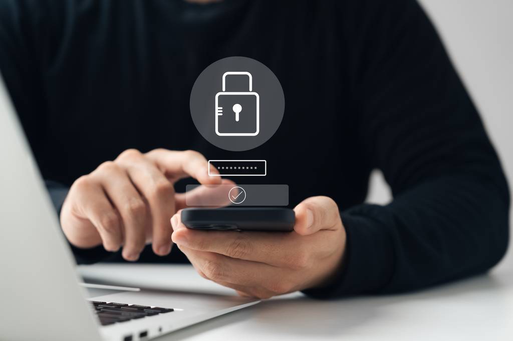 Ein Mann sitzt am Laptop und tippt auf seinem Smartphone ein Passwort ein.