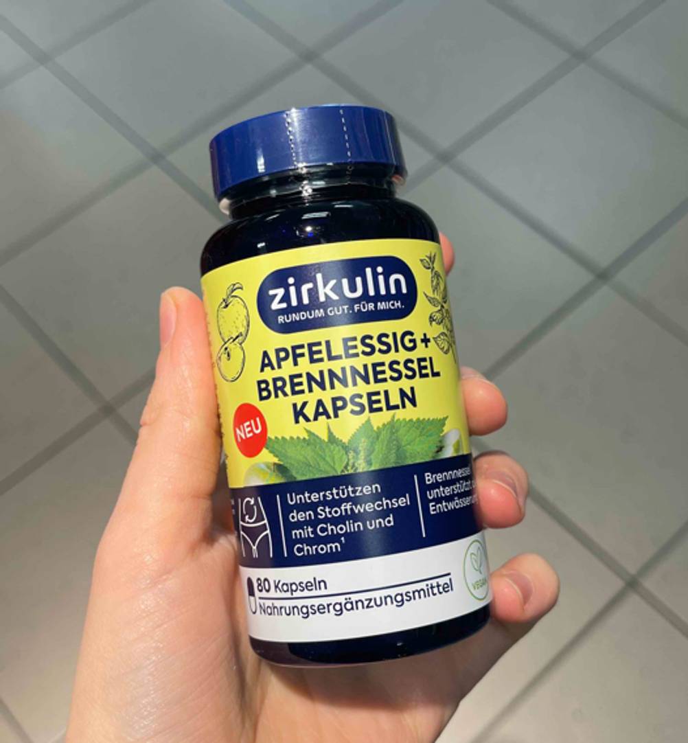 Apfelessig-Kapsel-Test: Eine Hand hält die blickdichte Kunststoffdose von Zirkulin vor einem grauen Fliesenhintergrund.