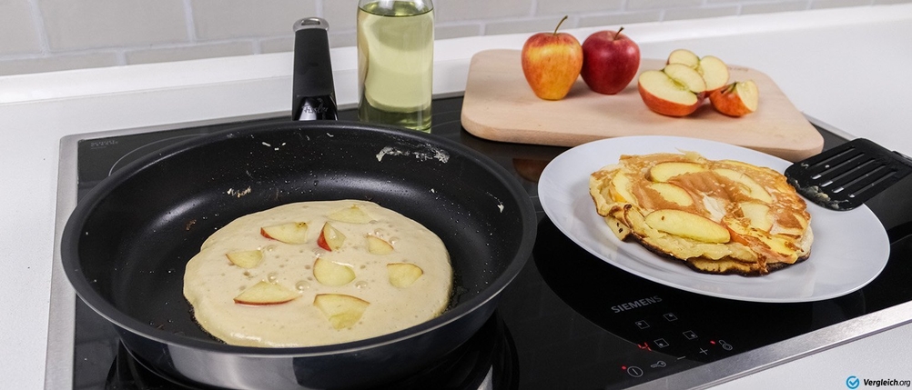 Pfannen-Test: In einer Pfanne wird ein Pfannenkuchen mit Apfelstücken gebacken, daneben steht ein Teller mit fertigen Pfannenkuchen sowie eine Schneidebrett mit Äpfeln.