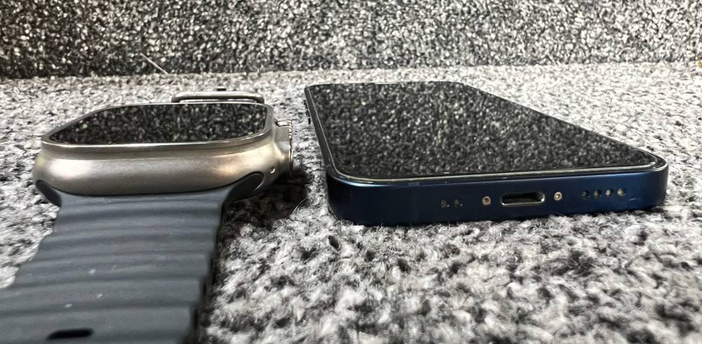 Nahaufnahme von einer grauen getesteten EKG-Smartwatch neben einem Smartphone auf schwarz-weiß meliertem Untergrund.