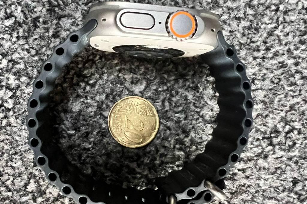 Eine Smartwatch-EKG zum testen liegt seitlich auf grauem Untergrund, in der Mitte ein 20-Cent-Stück.