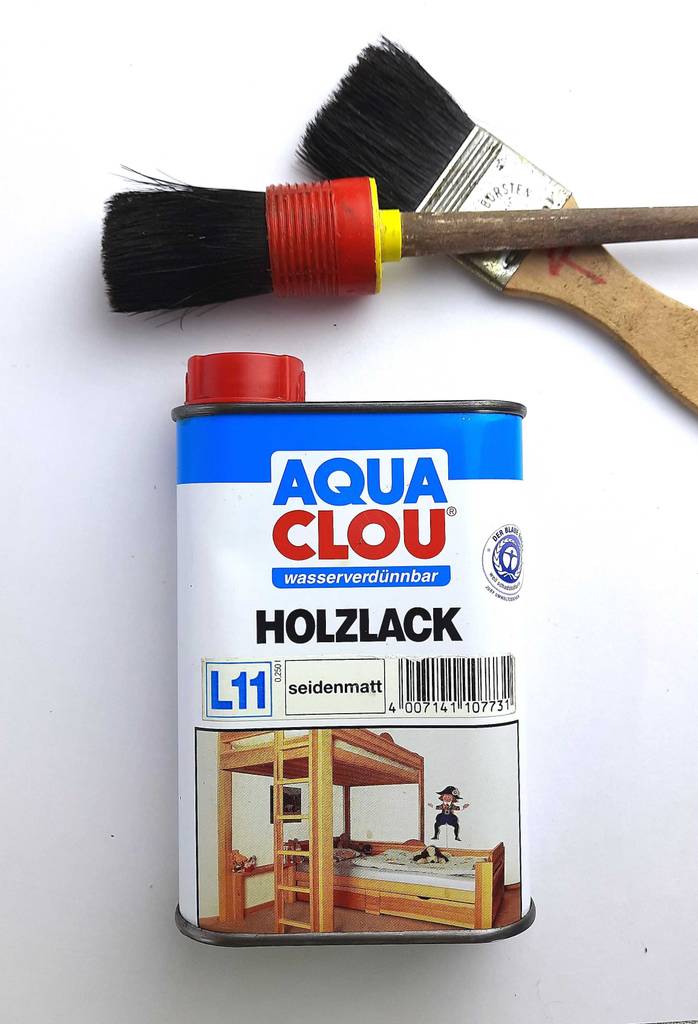 Holz-Klarlack-Test: Ein wasserverdünnbarer Holzlack liegt in seiner Dose unterhalb zweier Pinsel auf einer weißen Oberfläche.