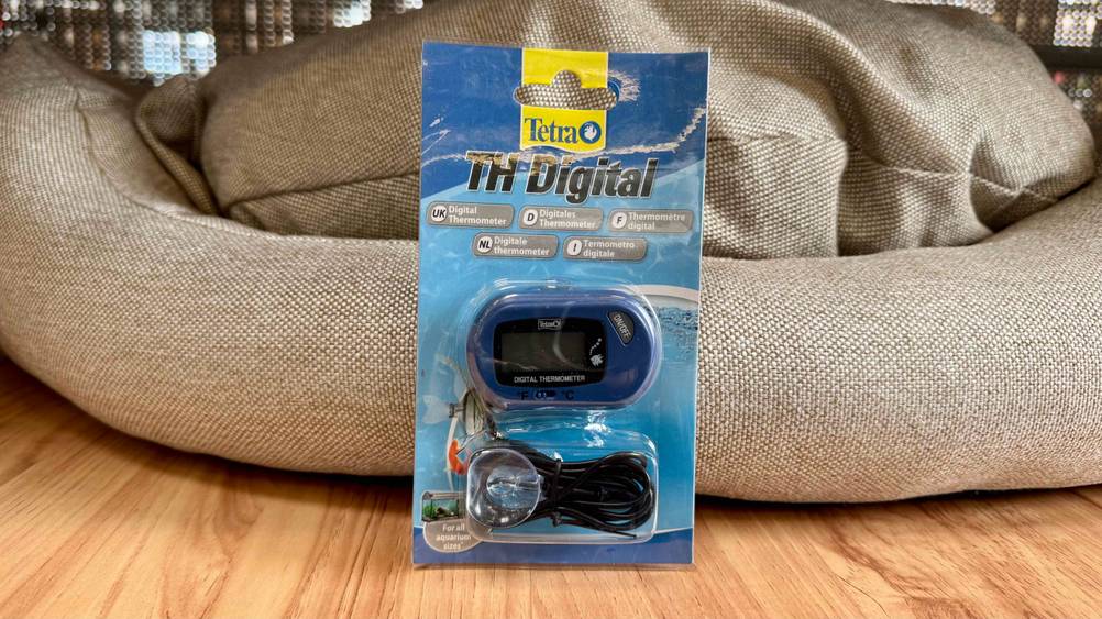 Ein getestetes Aquarium-Thermometer in transparenter Verpackung vor einem Kissen.