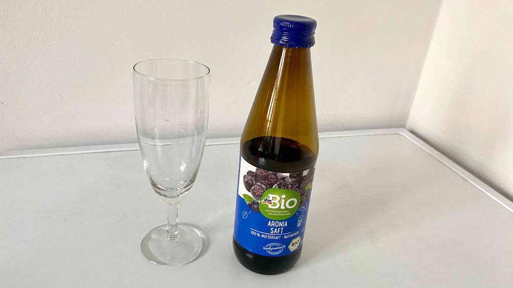 Eine Flasche des getesteten Aroniasaftes neben einem leeren Glas auf einer hellen Fläche.